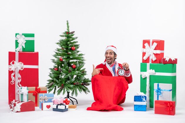 礼物微笑有趣的年轻人庆祝新年或圣诞节假期坐在地上 拿着礼物和装饰圣诞树附近的时钟微笑圣诞节抱着