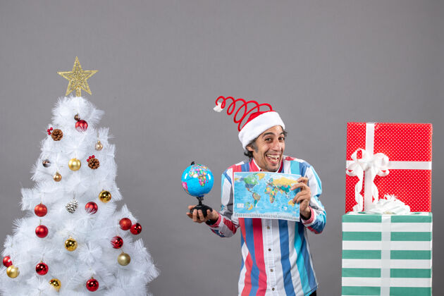喜出望外前视图欣喜若狂的男子与螺旋弹簧圣诞帽举行世界地图和地球仪周围圣诞树和礼物灰色男人印刷