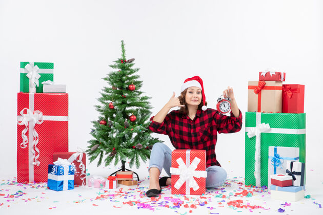 举行前视图的年轻女子坐在圣诞礼物周围拿着白色墙上的时钟节日雪坐