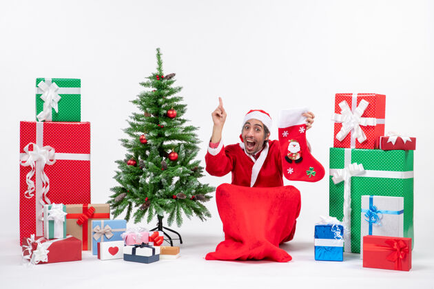礼物惊喜积极有趣的圣诞老人指着上面坐在地上 拿着圣诞袜靠近礼物和装饰在白色背景上的新年树正面袜子圣诞老人