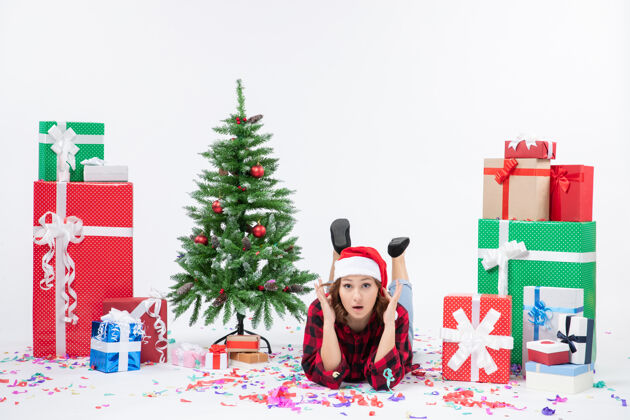 十二月年轻女子躺在白色墙壁上的圣诞礼物和小圣诞树周围的正视图人雪礼物