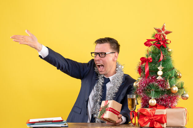 礼物一个年轻人对着坐在圣诞树旁的桌子上的人大喊大叫 并把礼物穿上黄色坐着年轻人商人