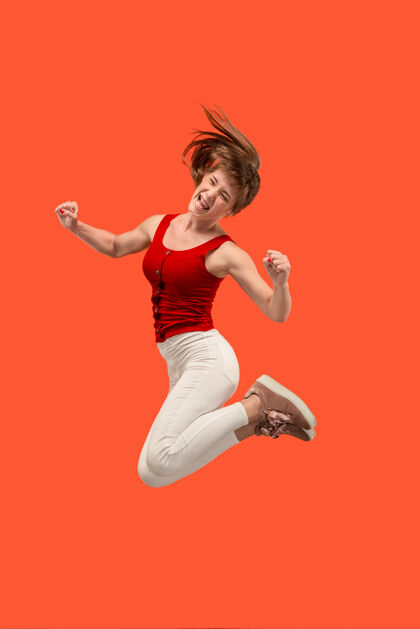 肖像移动中的自由在橙色工作室背景下 美丽快乐的年轻女子跳跃和做手势的半空中镜头在运动中奔跑的女孩人类情感和面部表情的概念无忧无虑表情冠军