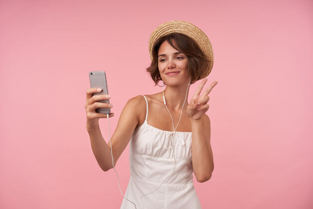智能手机快乐可爱的年轻女子 棕色短发 穿着夏装 戴着耳机 手持智能手机 用胜利手势举手自拍 与世隔绝皮带女士粉色