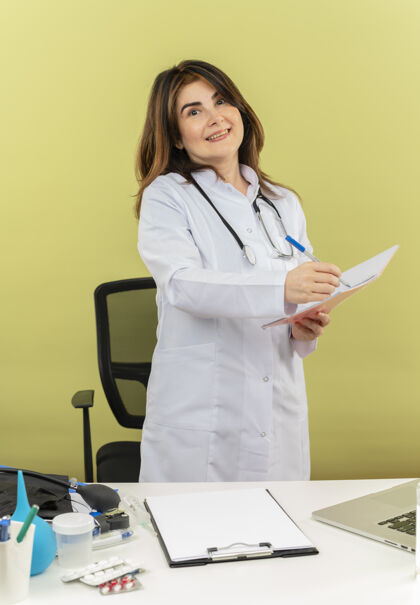 长袍微笑的中年女医生 穿着医用长袍 带听诊器 坐在办公桌旁 用笔记本电脑和医疗工具 拿着笔和笔记本 还有复印空间中年医疗笔记本电脑