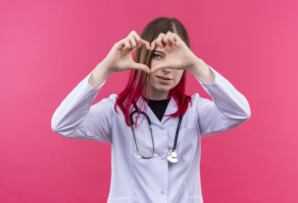 穿着穿着听诊器医用长袍的年轻医生女孩很高兴 在粉色孤立的背景上显示出心脏的姿态年轻礼服心脏