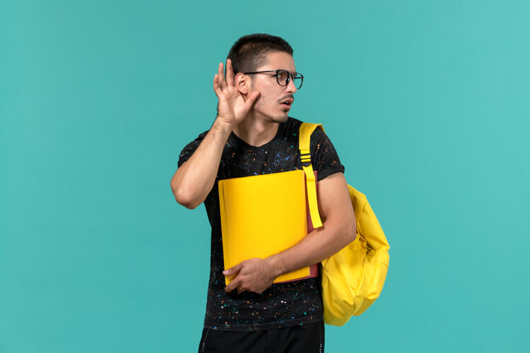 持有前视图中的男学生在深色t恤黄色背包举行不同的文件试图听到浅蓝色的墙上人男光