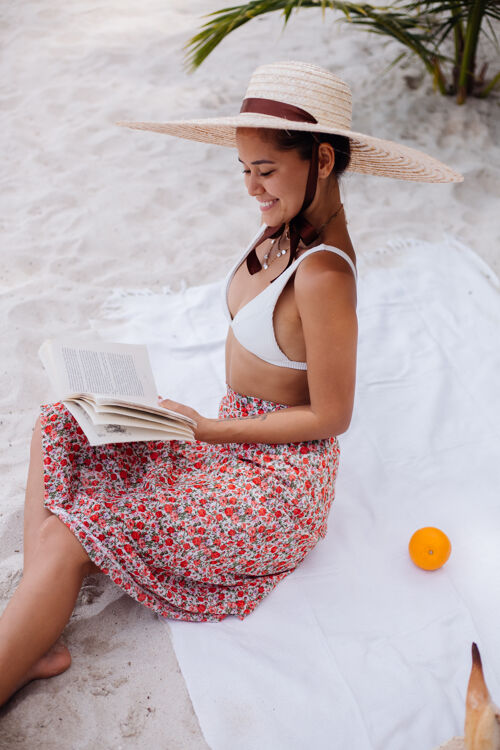 度假一个年轻女子戴着草帽 穿着白色针织衣服坐在沙滩地毯上自然女士海滩