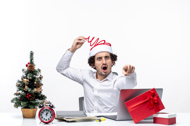 年轻的商人新的一年的心情与惊讶的年轻商人触摸他有趣的圣诞老人帽子指向在白色背景的办公室向前的东西微笑人办公室