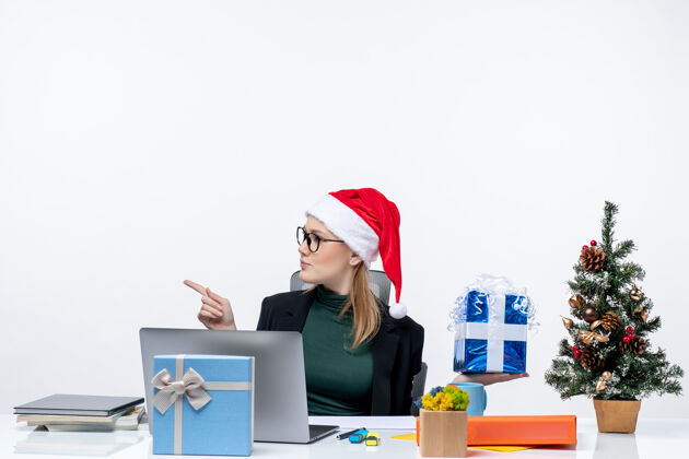 女人带着圣诞老人帽子戴着眼镜的年轻女子坐在桌子旁 展示着圣诞礼物 在白色背景上指着右边的东西 充满了好奇和惊讶的节日气氛笔记本电脑节日礼物