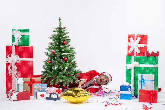 悲伤圣诞心情与年轻悲伤的圣诞老人躺在圣诞树后 在白色背景上不同颜色的礼物附近年轻圣诞树圣诞老人