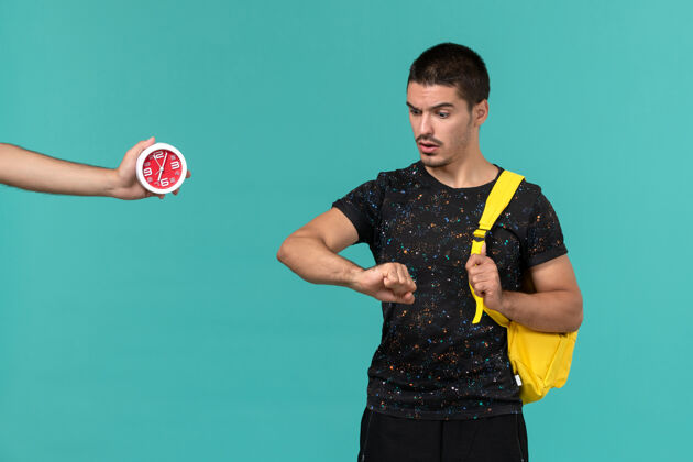 游戏身穿深色t恤黄色背包的男学生在浅蓝色的墙上看着他的手腕灯光学生男性