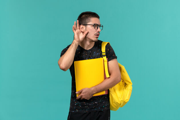 持有前视图中的男学生在深色t恤黄色背包举行不同的文件试图听到浅蓝色的墙上人男光