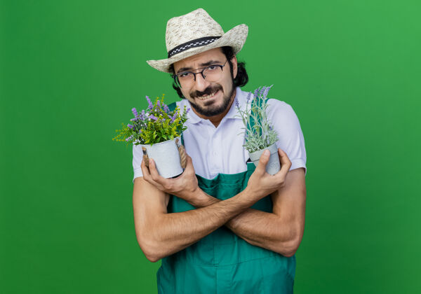 年轻年轻的留着胡子的园丁 穿着连体衣 戴着帽子 手里拿着盆栽植物 担心不高兴盆栽不高兴站着