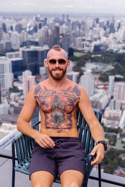 泰国成功的帅哥 留着胡子 野蛮的纹身 强壮的男人 赤裸上身 戴着手表 坐在高楼的椅子上 城市景色令人惊叹严肃人野蛮
