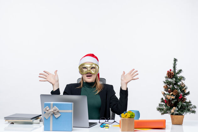办公室圣诞气氛与震惊的年轻女子戴着圣诞老人的帽子和面具坐在一张白色背景的桌子上圣诞老人工作人