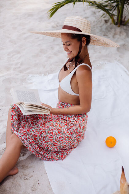 度假一个年轻女子戴着草帽 穿着白色针织衣服坐在沙滩地毯上自然女士海滩
