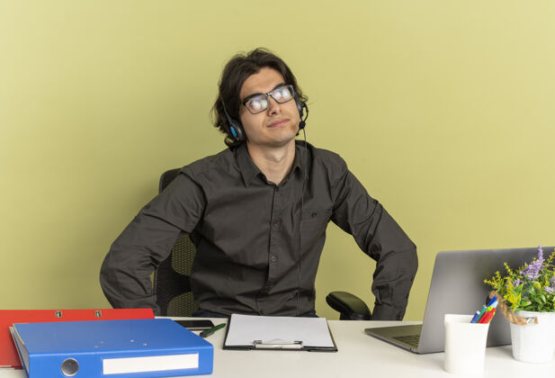 坐年轻体贴的上班族戴着耳机 戴着眼镜 坐在办公桌旁 用笔记本电脑拿着办公工具 看着旁边看光学年轻
