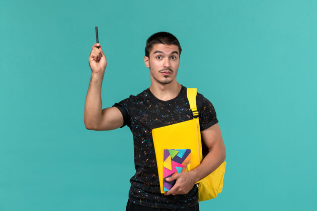 男性前视图中的男学生穿着深色t恤黄色背包拿着文案笔和文件在浅蓝色的墙上人笔正面