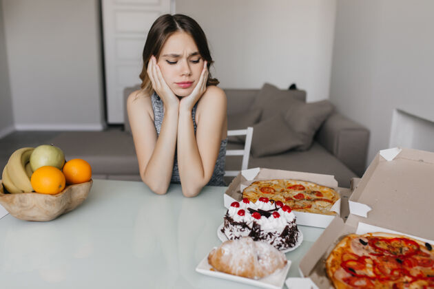 金发悲伤的卷发女人在节食时看着蛋糕金发美女模特摆着水果和比萨饼的姿势巧克力快餐选择