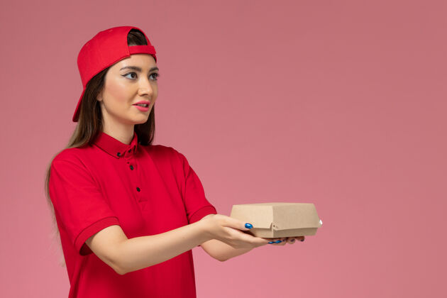 年轻正面图：身穿红色制服和斗篷的年轻女快递员 手上拿着一个小小的快递食品包 放在粉红色的墙上手快递女性
