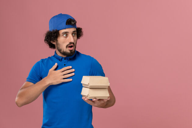 查看正面图：身穿蓝色制服 披风的男性快递员在粉红色的墙上拿着小包裹男性快递粉色送货