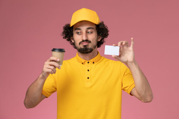 送货正面图：身穿黄色制服 披风的男性快递员手持咖啡杯和粉色墙上的卡片男快递粉色工人