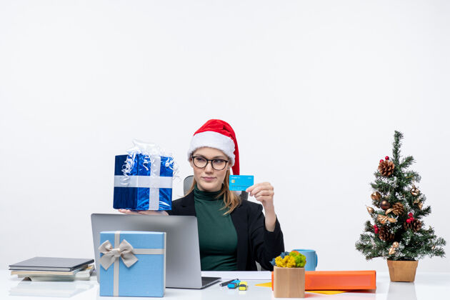 商务一位戴着圣诞老人帽 戴着眼镜 拿着圣诞礼物和白色背景的银行卡坐在桌旁的商务女士大吃一惊眼镜女士拿着