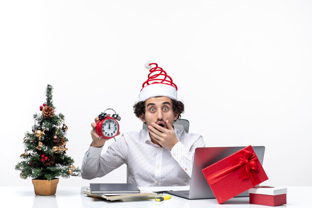 独自年轻兴奋震惊的商人戴着圣诞老人的帽子 在白色的背景下独自一人坐在办公室里看时钟圣诞老人人生活