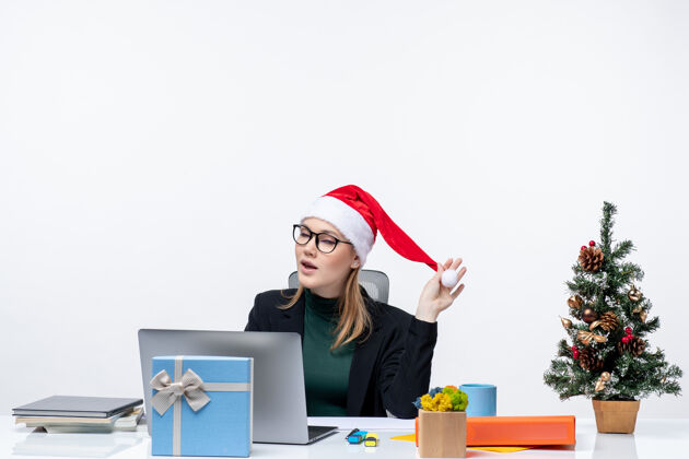 桌子情绪激动的女商人坐在一张桌子旁 手里拿着圣诞树和礼物 手里拿着一顶圣诞老人的帽子 在白色背景下查看邮件办公室圣诞老人情绪化