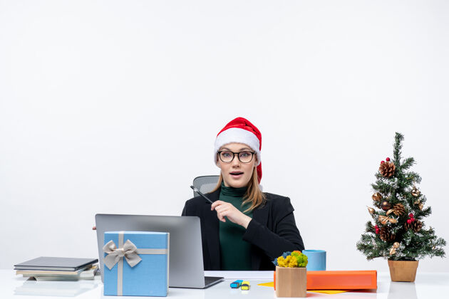 礼物一个戴着圣诞老人帽子的金发女人坐在一张桌子旁 桌子上有一棵圣诞树和一份礼物 在白色背景上指着右边的东西圣诞节桌子坐着