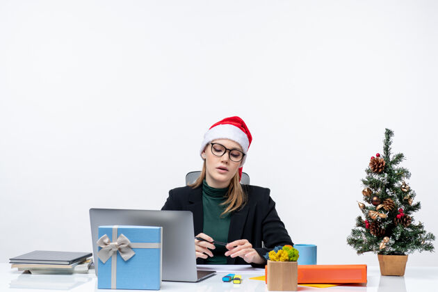 小心困惑的金发女人 戴着圣诞老人的帽子 坐在一张桌子旁 桌子上放着圣诞树和礼物 在白色背景下仔细地看着办公室里的东西桌子帽子工作