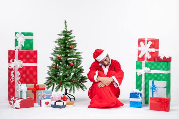 年轻悲伤不快乐的年轻人打扮成圣诞老人 拿着礼物和装饰过的圣诞树坐在白色背景的地上礼物圣诞树圣诞老人