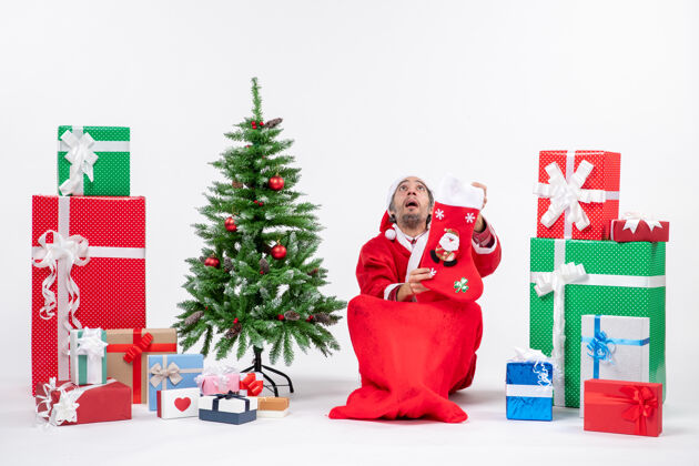 地悲伤的圣诞老人看着上面坐在地上 拿着圣诞袜靠近礼物和装饰在白色背景上的新年树坐着圣诞圣诞老人