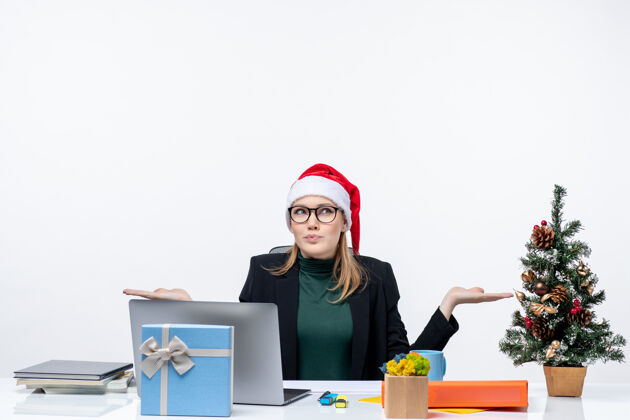 礼物积极好奇的女商人 戴着圣诞老人帽 坐在一张桌子旁 桌子上放着圣诞树和一件白色背景的礼物电脑商务女人人