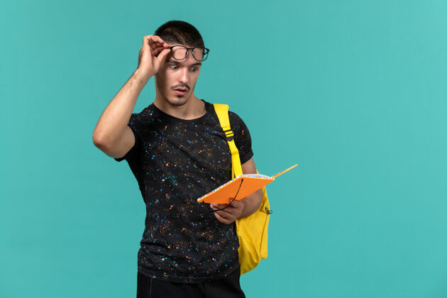 冒充身穿深色t恤黄色背包的男学生的正面图拿着蓝色墙壁上的抄写本阅读字帖男学生肖像