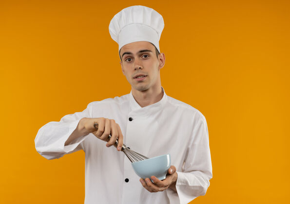 穿年轻男性穿着厨师制服 拿着搅拌器和碗站在隔离的黄色墙上年轻酷男