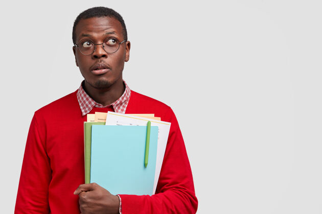 思考黑人学生带着沉思的表情向上看 拿着课本 戴着眼镜以获得良好的视力体贴教科书空白