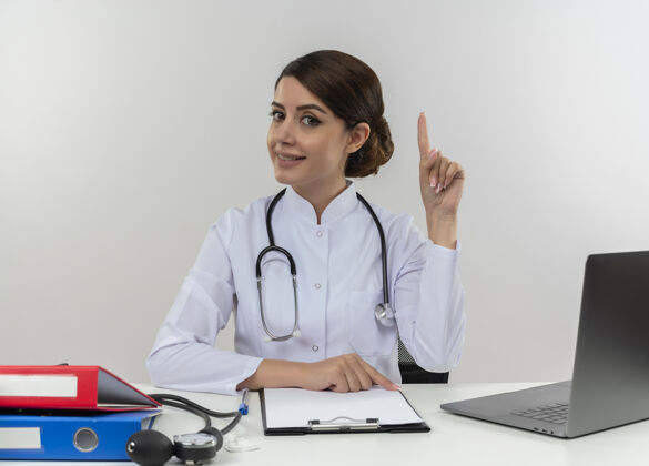 医生高兴的年轻女医生穿着医用长袍拿着听诊器坐在办公桌上用电脑拿着医疗工具指着上面的复印空间穿听诊器工作