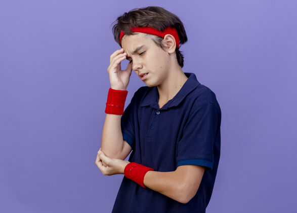 头带疼痛的年轻英俊的运动男孩戴着头带和腕带触摸头部保持手肘向下看患有头痛孤立触摸手头痛