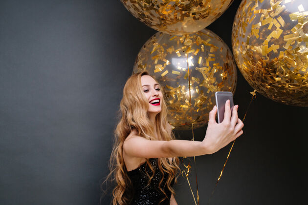 快乐穿着黑色豪华连衣裙的年轻漂亮女人的现代派对时间 长卷曲的金发与装满金色金属片的大气球自拍庆祝 微笑金箔明亮除夕夜