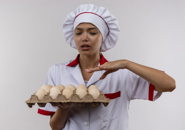 鸡蛋困惑的年轻厨师女身穿厨师制服拿着一批鸡蛋站在隔离的白色墙上厨师女持有