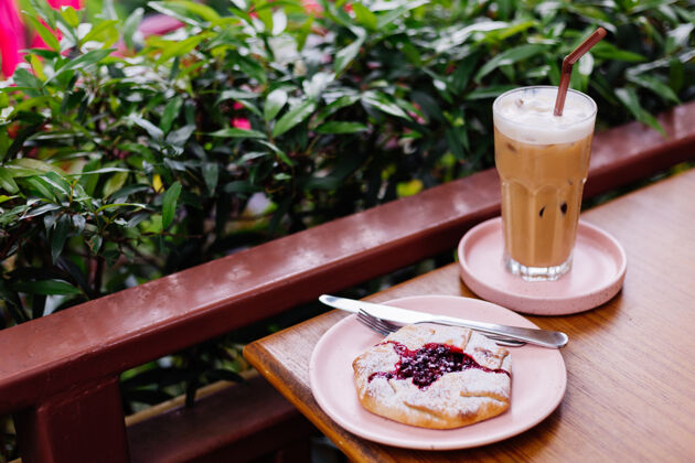 清凉木制桌子上粉红色的架子上放着冰拿铁 夏天咖啡馆绿树丛中放着小红莓馅饼卡路里棕色咖啡馆