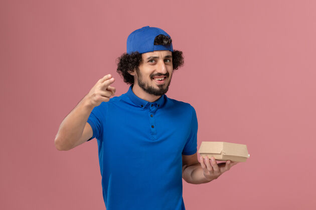 男性快递正面图身穿蓝色制服和披风的男性快递员拿着粉色墙上的小快递食品包制服人肖像