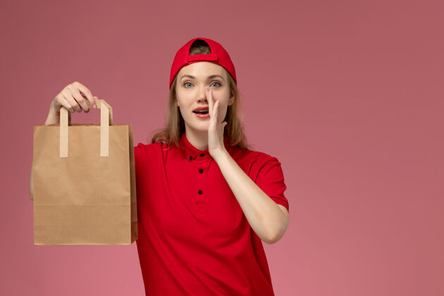 呼叫正面图：身穿红色制服 披风的年轻女快递员手持快递食品包 在粉色墙上呼喊手持人漂亮