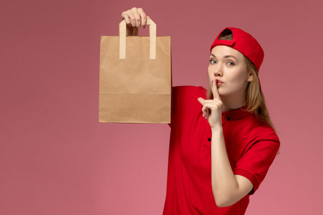 人正面图：身穿红色制服 披风的年轻女快递员拿着快递食品包站在浅粉色的墙上微笑快递快乐