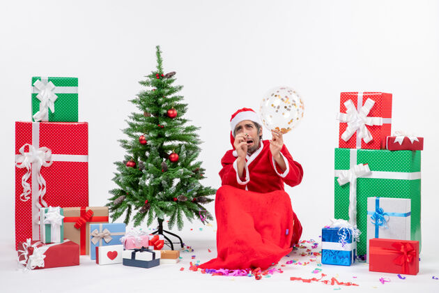 害怕圣诞气氛与恐惧的圣诞老人举行气球坐在圣诞树和礼物在白色背景上不同的颜色气球抱着人