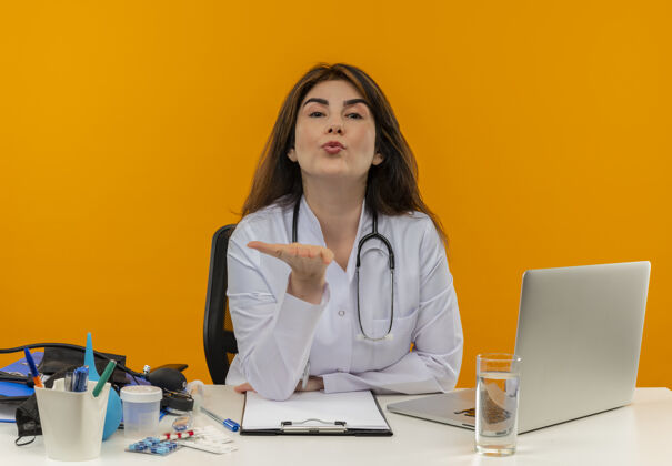 笔记本电脑中年女医生穿着医用长袍 带听诊器 坐在办公桌旁 用笔记本电脑和医疗工具 在隔离的橙色墙壁上显示亲吻手势 并留有复印空间办公桌中年医疗